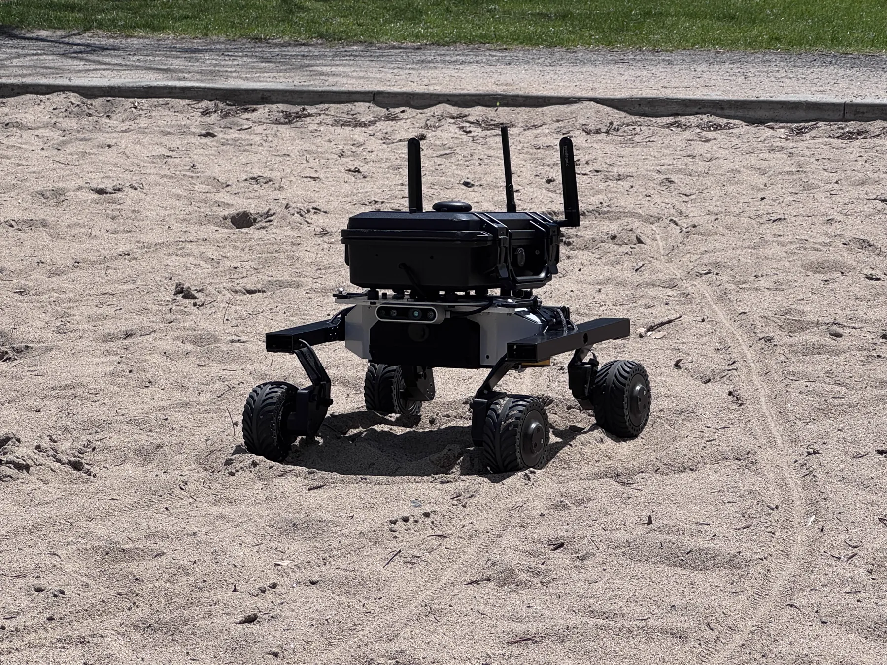 A rugged UGV carrying a sensor payload while driving through a sandy field