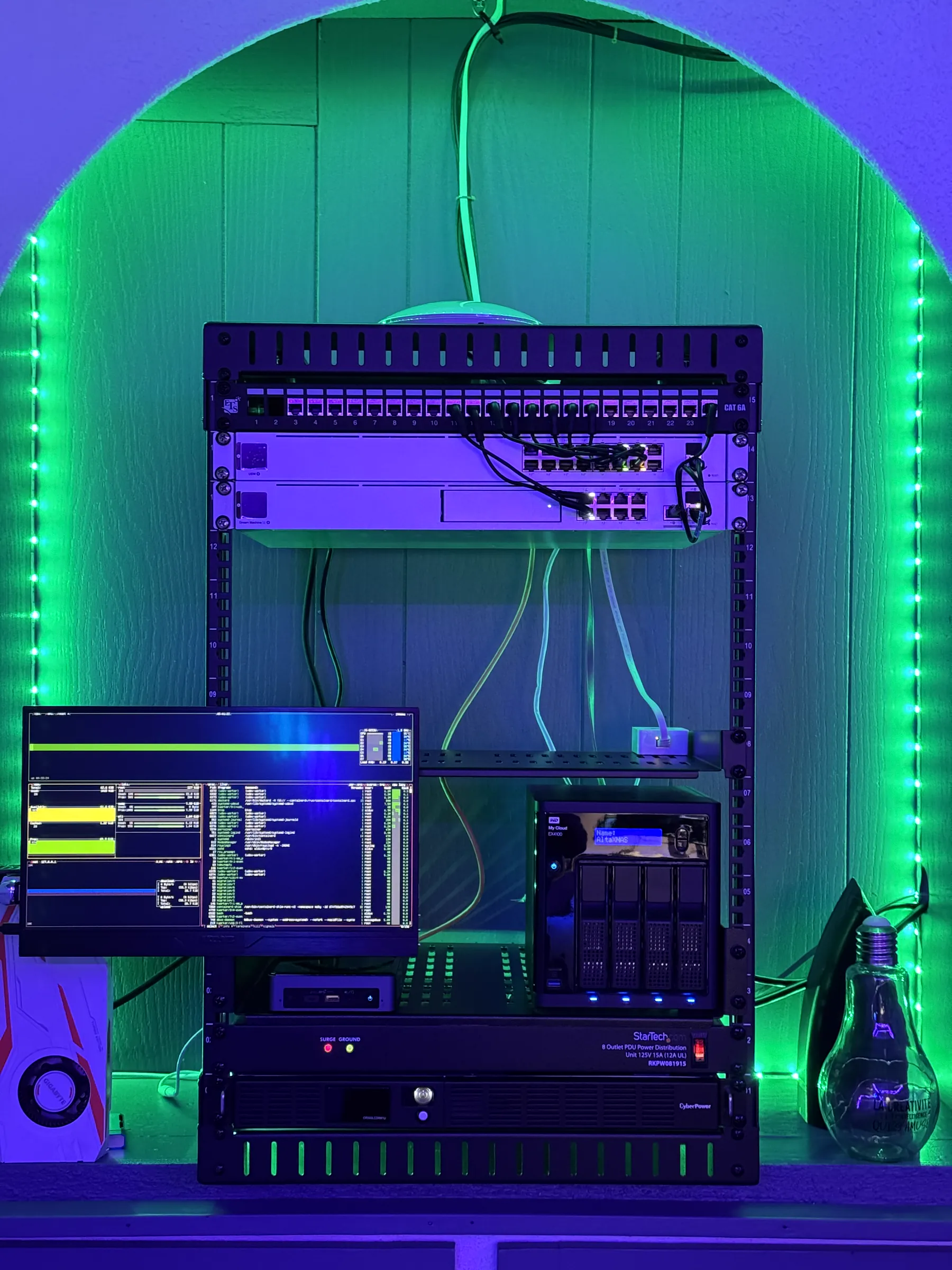 Green-lit home-lab server and network rack with switches, a NAS labelled AltaNAS and a monitor running a monitoring dashboard